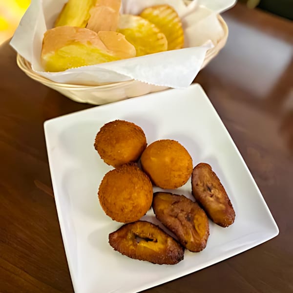 A white plate of golden-brown fried plantains and croquettes, with a basket of pastries in the background.