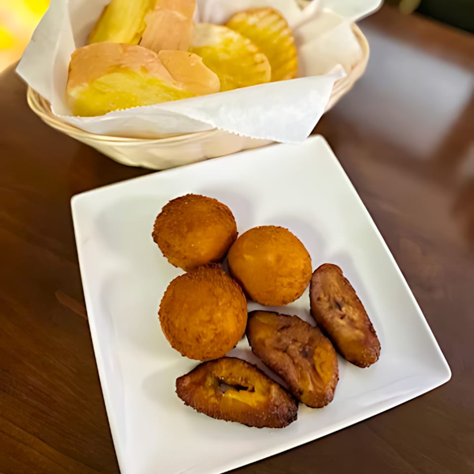 A white plate of golden-brown fried plantains and croquettes, with a basket of pastries in the background.
