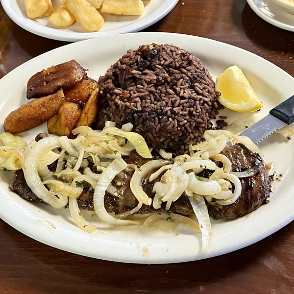 Cuban steak with onions, black beans and rice, fried plantains, and a lemon wedge.