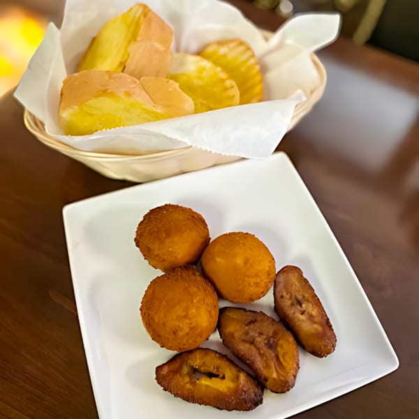 A plate of fried plantains and croquettes, with a basket of bread in the background.